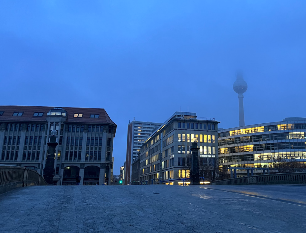 Looking towards the building of the Theologische Fakultät on the left, a grey building of five floors with a sharply sloped red roof, with other nearby buildings to the right of a similar height, and the tall Fernsehturm in the background towering over all, and disappearing at its pointy top into the fog