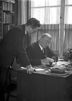 Auf dem Bild sieht man Karl Themel. Er sitzt an einem Tisch und schreibt. Neben ihm steht Horst Schirmacher. Das Foto entstand im Jahr 1933.