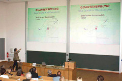 Blick über die Köpfe des Publikums im großen Hörsaal. An die Wand sind Präsentationsfolien projeziert, auf ihnen steht "Quantensprung".